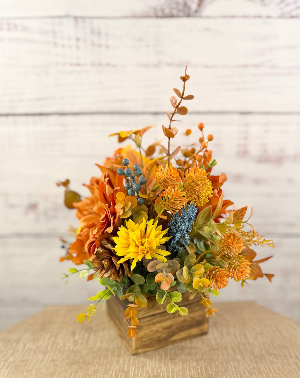 Cozy Harvest fall arrangement with orange and yellow flowers, blue accents and pinecones in a rustic wooden cube