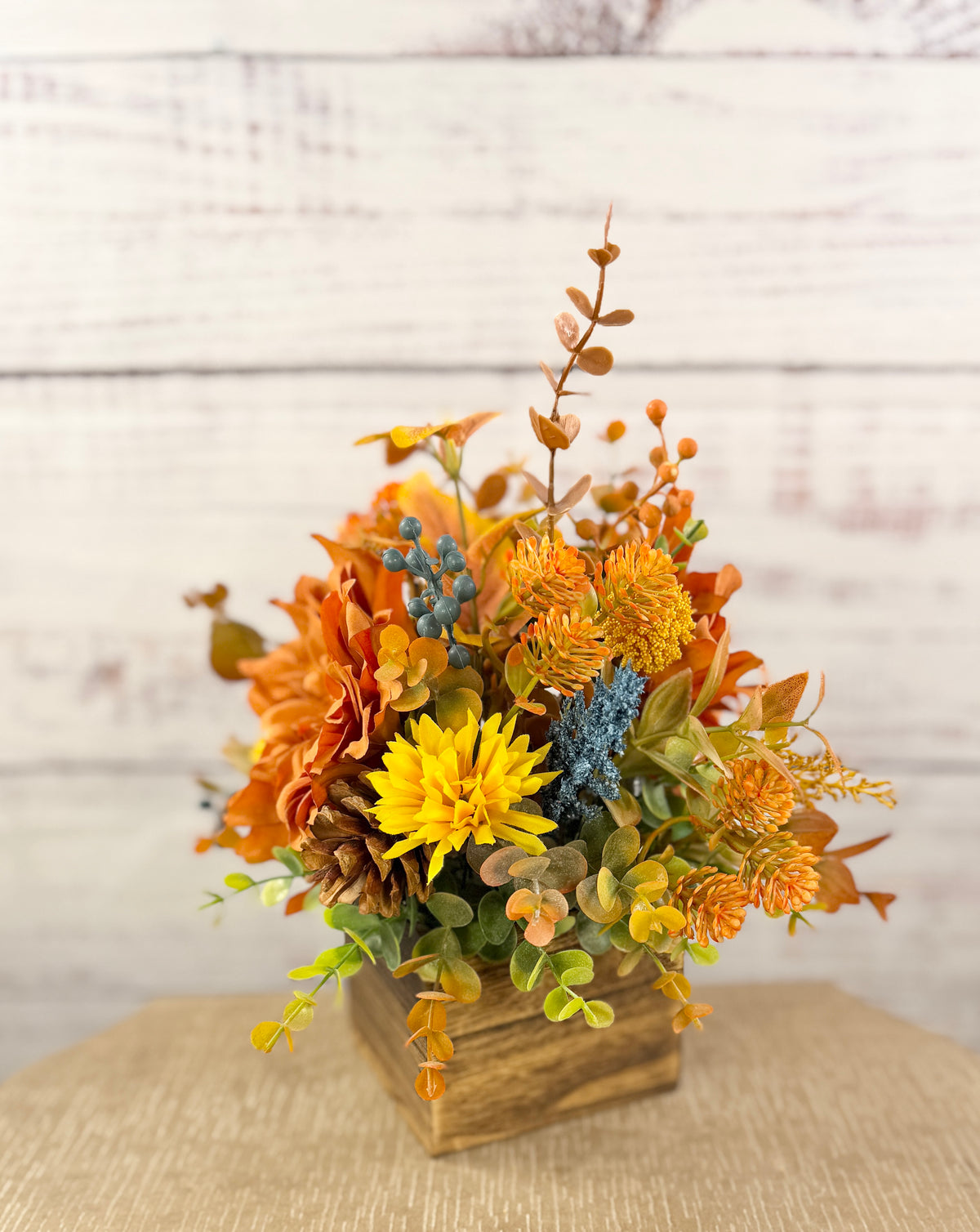 Cozy Harvest fall arrangement with orange and yellow flowers, blue accents and pinecones in a rustic wooden cube