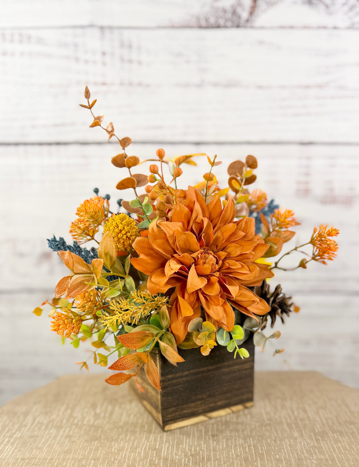Cozy Harvest fall arrangement with orange and yellow flowers, blue accents and pinecones in a rustic wooden cube