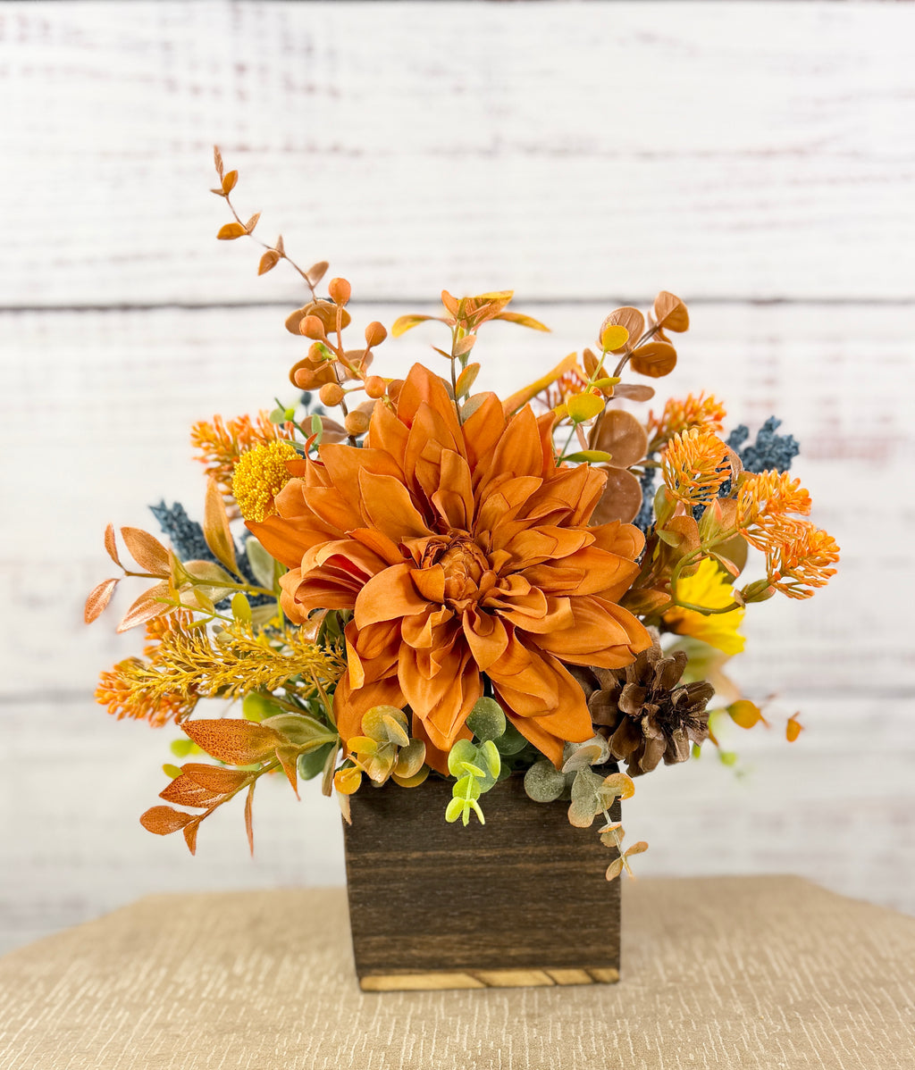 Cozy Harvest fall arrangement with orange and yellow flowers, blue accents and pinecones in a rustic wooden cube