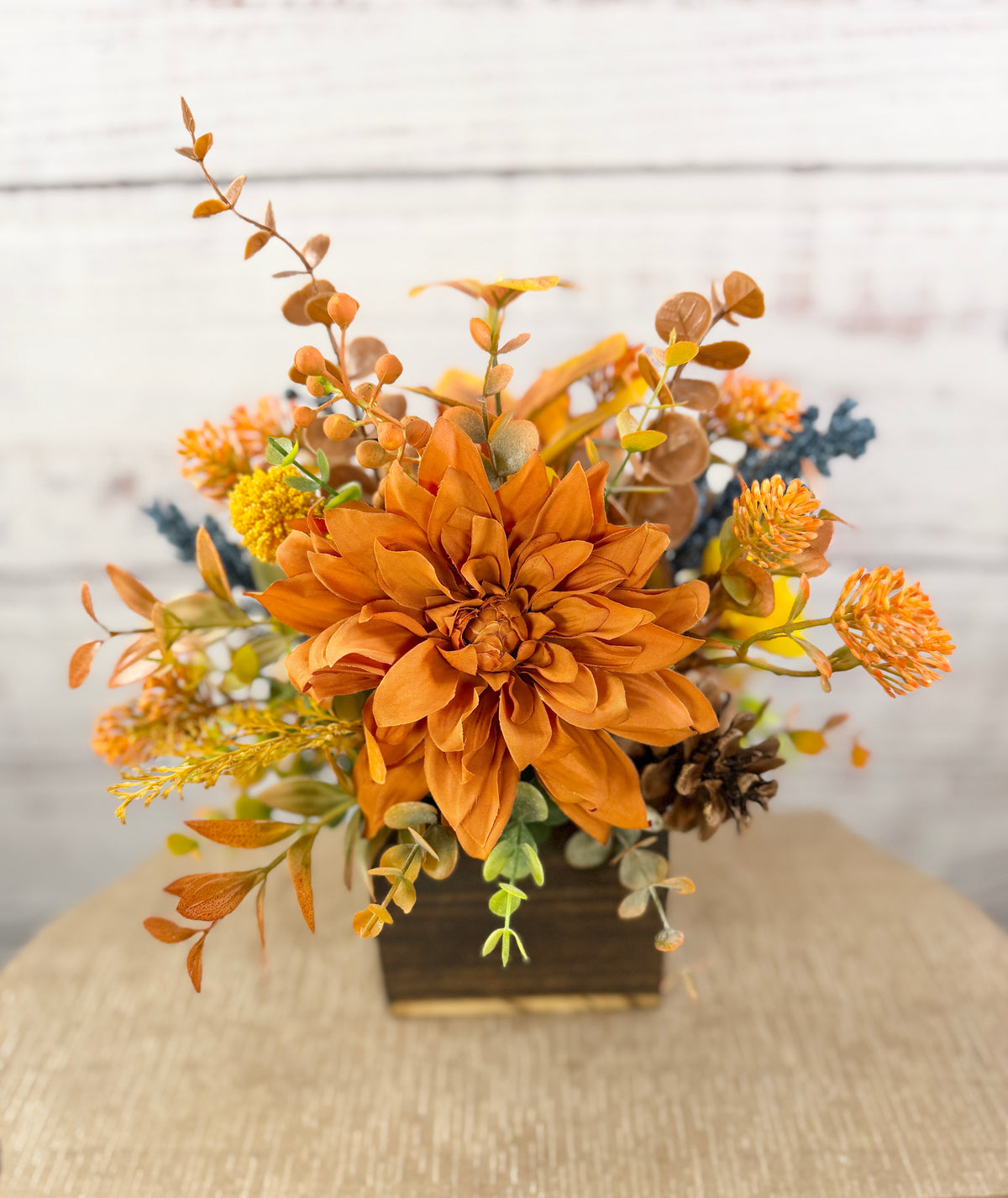 Cozy Harvest fall arrangement with orange and yellow flowers, blue accents and pinecones in a rustic wooden cube