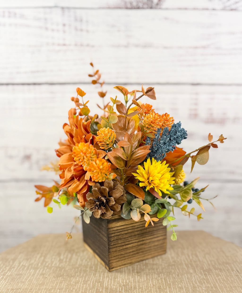 Cozy Harvest fall arrangement with orange and yellow flowers, blue accents and pinecones in a rustic wooden cube
