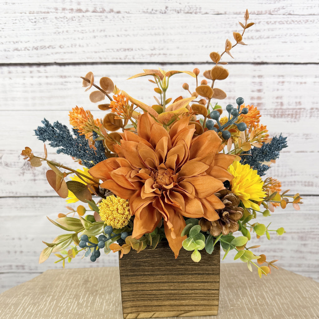 Cozy Harvest fall arrangement with orange and yellow flowers, blue accents and pinecones in a rustic wooden cube